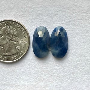 May include: Two oval, faceted blue gemstones next to a U.S. quarter. The gemstones are a deep blue with lighter streaks. The quarter shows George Washington's profile. The gemstones are approximately 1.5 cm in length.