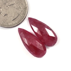 Puede incluir: Dos gemas facetadas, en forma de lágrima, de color rojo intenso se muestran sobre una superficie blanca junto a una moneda. Las gemas tienen una superficie pulida y reflectante.