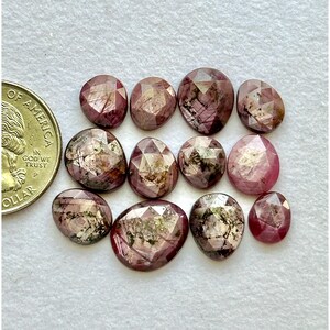 May include: A collection of faceted, oval-shaped gemstones in shades of pink and burgundy. The gemstones are arranged on a white surface next to a US quarter coin. The gems have a polished, reflective surface.