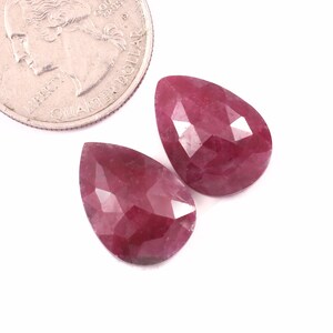 May include: Two faceted, pear-shaped, deep red gemstones are displayed next to a quarter coin. The gemstones have a polished, reflective surface and are set against a white background. The gems are likely rubies.