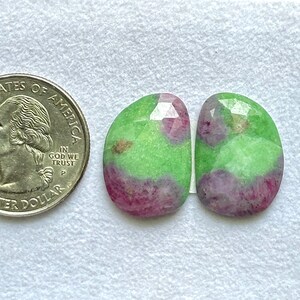 Puede incluir: Dos piedras preciosas pulidas con una moneda de veinticinco centavos para comparar el tamaño. Las piedras preciosas tienen forma ovalada, con un color verde vibrante y rojo intenso. Superficie facetada.