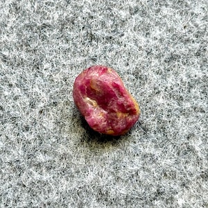 May include: A rough, natural ruby gemstone. The ruby is a deep pinkish-red color with some lighter, yellowish areas. The stone has an irregular shape and a slightly textured surface. It is set against a gray, textured background.