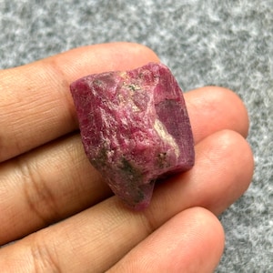May include: A raw ruby gemstone, displaying a rich red colour. The surface has a textured appearance with lighter and darker patches. The gemstone is held in a hand against a grey background.