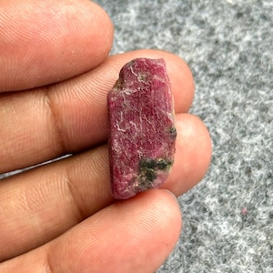 May include: A rough-cut ruby gemstone, displaying a deep red hue with areas of darker coloration and some green inclusions. The raw mineral is held in a person's hand, showcasing its natural texture and form.