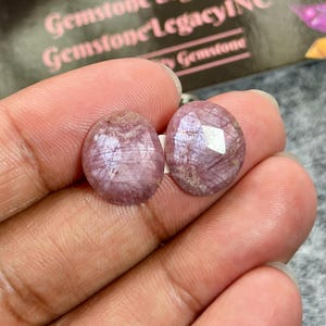 May include: Two round, faceted, translucent pinkish-purple gemstones with visible internal inclusions and a polished surface. The stones exhibit a rose-toned hue with subtle variations in color and light reflection across their cut, geometric facets.