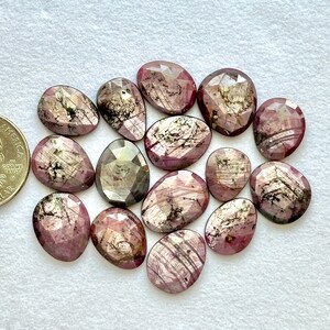 May include: A collection of faceted, oval-shaped gemstones in shades of pink and gray. The gemstones have internal patterns and are arranged on a white surface. The gems vary in size and shape, with a coin for scale.