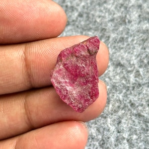 Peut inclure: Une pierre précieuse brute de rubis non taillée, d'un rouge profond. Le minéral brut présente une surface texturée et une forme irrégulière, révélant des formations naturelles. Le rubis est tenu entre deux doigts, soulignant sa taille et sa couleur.