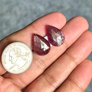 May include: Two faceted, teardrop-shaped, deep red gemstones are displayed next to a quarter coin. The gemstones have a sparkling, geometric cut, and are set against a neutral background. The gemstones are likely rubies.