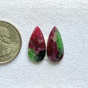 May include: Two pear-shaped faceted gemstones featuring a mix of deep red, vibrant green, and black inclusions. The stones are positioned next to a United States one dollar coin for scale, showing the relative size of the polished mineral pieces.