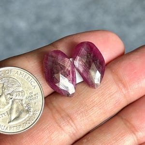 Peut inclure: Deux pierres précieuses rubis en forme de poire à facettes, aux teintes magenta et rouge profond, présentant des inclusions naturelles et des surfaces réfléchissantes, placées à côté d'une pièce de 25 cents américaine pour l'échelle.