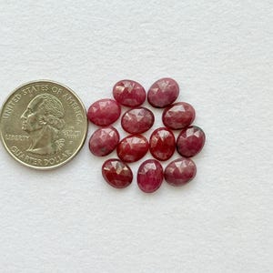 Peut inclure: Un ensemble de pierres précieuses ovales, facettées, rouge foncé. Une pièce de monnaie américaine de 25 cents est placée à côté des pierres précieuses pour la comparaison des tailles. Les pierres précieuses sont disposées sur une surface blanche.