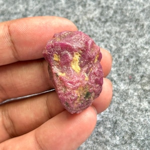 Puede incluir: Una gema de rubí en bruto sin cortar. El rubí es de color rojo intenso con áreas amarillas y verdes. La piedra tiene una superficie texturizada y se sostiene en una mano.