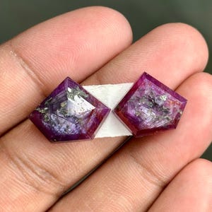 Pode incluir: Dois cabochões de pedra preciosa roxa e vermelha com um corte facetado em forma de pipa. As pedras têm um aspecto salpicado com manchas prateadas.