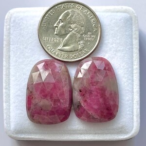 Könnte beinhalten: Zwei facettierte, oval geformte rosa Edelsteine mit einer Vierteldollarmünze zum Größenvergleich. Die Edelsteine haben eine Mischung aus hellen und dunklen Rosatönen und liegen in einer weißen Box.