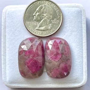 Könnte beinhalten: Zwei facettierte, oval geformte Edelsteine mit rosa und grauen Farbtönen. Eine Vierteldollarmünze liegt zur Größenveranschaulichung darüber. Die Edelsteine liegen auf einem weißen Hintergrund.