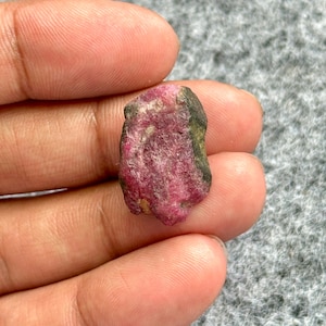 Peut inclure: Une pierre précieuse de rubis naturelle, taillée grossièrement. Le rubis est d'un rouge profond, avec des zones plus foncées et quelques inclusions vertes et brunes. La pierre est tenue dans une main, montrant sa forme irrégulière et sa texture naturelle.