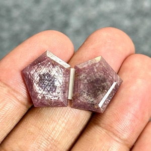 Puede incluir: Dos rubíes facetados en forma de pentágono, de un tono rojo rosáceo intenso con inclusiones internas visibles. Las piedras preciosas presentan una superficie superior plana con bordes angulares, una textura natural semitranslúcida y un acabado pulido.