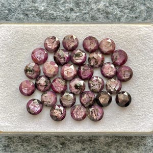 Peut inclure: Une collection de vingt-cinq pierres précieuses rondes et facettées dans des tons rose foncé, magenta et rouge sombre. Chaque pierre présente une taille rose avec des inclusions internes visibles, des variations naturelles et une surface polie.