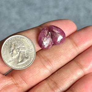 Puede incluir: Dos gemas facetadas, en forma de lágrima, de color rosa-rojo. Una moneda de veinticinco centavos estadounidense se encuentra junto a las gemas para comparar el tamaño. Las gemas tienen una calidad translúcida y están sobre un fondo neutro.