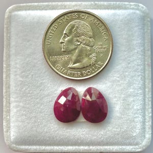 May include: Two faceted, teardrop-shaped, deep red rubies are displayed next to a U.S. quarter coin in a white velvet-lined box. The coin shows a profile of George Washington and the words "United States of America".