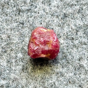 Puede incluir: Una piedra preciosa de rubí rojo intenso con una superficie rugosa y natural. El rubí es de un color rojo vibrante y tiene una forma ligeramente irregular.