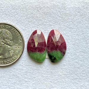 Puede incluir: Dos piedras preciosas de rubí zoisita talladas en forma de pera, con una clara división de color entre el rojo magenta intenso y el verde musgo, colocadas junto a una moneda de cuarto de dólar estadounidense para mostrar su tamaño.