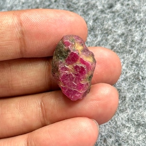 May include: A rough, uncut ruby gemstone with a deep red hue, speckled with areas of green and yellow. The natural stone has an irregular shape and a slightly textured surface, showcasing its raw, unpolished state.