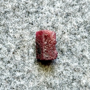 May include: A deep red ruby gemstone, cut into a rectangular shape, sits on a white, textured surface.