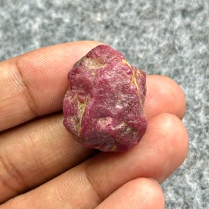May include: A rough, uncut ruby gemstone, showcasing a deep red hue with natural textures and slight variations in color. The raw mineral appears to be held in a hand, highlighting its natural form.