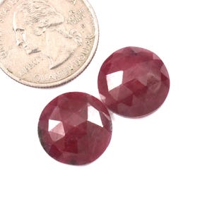 May include: Two round, faceted, deep pinkish-red gemstones positioned next to a United States quarter coin for scale. The stones feature a rose-cut style with multiple triangular facets reflecting light across their polished, circular surfaces.