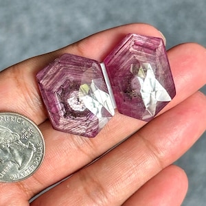 May include: Two faceted, hexagonal-shaped pinkish-red gemstones with visible internal inclusions and a polished surface. A United States quarter coin is positioned next to the stones to provide a clear sense of scale for the objects.