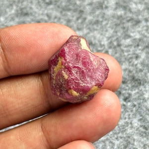 May include: A rough, uncut ruby gemstone with a deep red hue and patches of yellow and white. The natural stone has an irregular shape and a slightly textured surface. This raw ruby is a beautiful example of a natural gemstone.