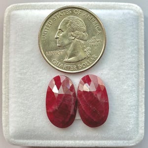 Puede incluir: Dos gemas facetadas, de forma ovalada y de color rojo intenso, se muestran junto a una moneda de veinticinco centavos estadounidense. Las gemas están colocadas en una caja forrada de terciopelo blanco. La moneda muestra un perfil de George Washington.