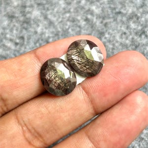 Op de afbeelding: Twee gefacetteerde, ovaalvormige edelstenen met een donkere, iriserende uitstraling. De stenen hebben een mix van bruine, grijze en zilveren tinten, met een sprankelend, reflecterend oppervlak. De edelstenen worden gepresenteerd op een lichtgrijze achtergrond.