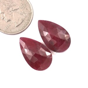 Puede incluir: Dos gemas facetadas, en forma de pera, de color rojo intenso se muestran junto a una moneda de veinticinco centavos estadounidense para comparar el tamaño. Las gemas tienen una superficie pulida y reflectante, sobre un fondo blanco.