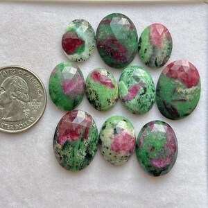 Puede incluir: Una colección de piedras preciosas ovaladas con superficies facetadas. Las piedras muestran una mezcla de tonos verdes y rojos, con una moneda para comparar el tamaño. Las piedras preciosas están dispuestas sobre una superficie blanca.