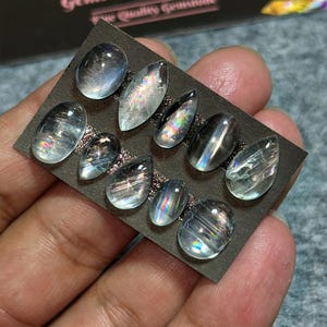 May include: A collection of ten iridescent gemstones in various shapes, including oval and teardrop, are displayed on a dark gray card. The stones have a translucent quality with internal rainbow-like flashes. The card is held by a hand.