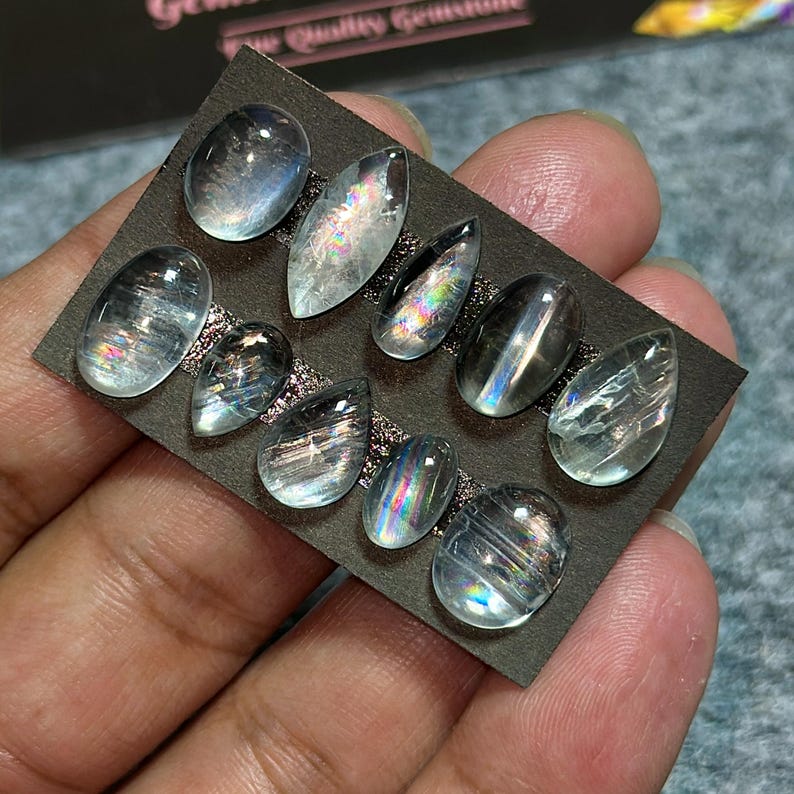 May include: A collection of ten iridescent gemstones in various shapes, including oval and teardrop, are displayed on a dark gray card. The stones have a translucent quality with internal rainbow-like flashes. The card is held by a hand.