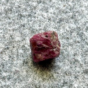 May include: A rough, uncut ruby gemstone with a deep red hue. The natural stone has a textured surface with areas of lighter pink and gray. The ruby is displayed on a gray, textured background.