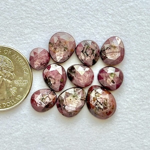 May include: A collection of faceted, oval-shaped gemstones in varying sizes. The gems are a deep pinkish-red color with internal inclusions. A US quarter is included for size reference. The gemstones are arranged on a white surface.