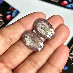 Pode incluir: Duas pedras preciosas ovais com uma tonalidade roxa e um acabamento brilhante e iridescente. As pedras têm um corte facetado e são exibidas em uma mão.