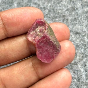 Peut inclure: Une pierre précieuse de rubis brute, présentant une teinte rouge profond avec des zones roses et vertes plus claires. La pierre naturelle a une surface texturée et une forme irrégulière, idéale pour la joaillerie ou la collection.