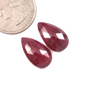 Puede incluir: Dos rubíes tallados en forma de pera con un color rojo intenso y superficies reflectantes, colocados junto a una moneda de cuarto de dólar estadounidense para comparar, mostrando el tamaño relativo de las gemas frente a los 24,26 mm de diámetro.