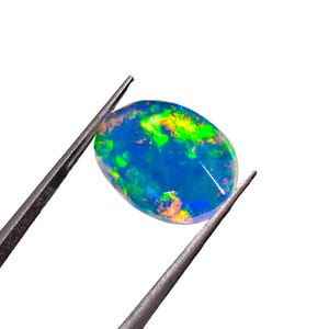 May include: An oval-shaped loose gemstone with a dark blue base color and vibrant flashes of neon green, yellow, and orange play-of-color. The stone is held by a pair of dark metal tweezers against a plain white background.