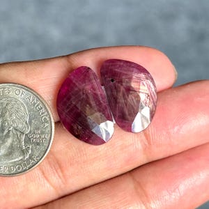 May include: Two faceted, teardrop-shaped deep magenta gemstones with visible internal inclusions and natural striations. A silver-colored United States quarter coin is positioned next to the stones for scale, showing their relative size.