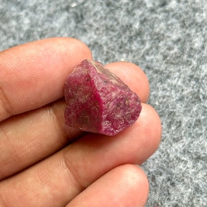Könnte beinhalten: Ein rauer, ungeschliffener Rubin-Edelstein mit tiefroter Farbe. Der Naturstein hat eine strukturierte Oberfläche und eine unregelmäßige Form, die seinen rohen, unpolierten Zustand zeigt. Der Rubin wird in einer Hand gehalten, mit grauem Hintergrund.