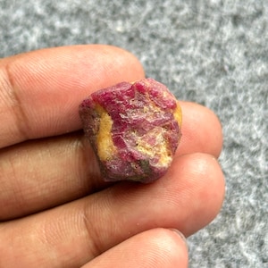 May include: A rough, uncut ruby gemstone with a deep red hue, accented by patches of yellow and tan. The natural stone has a textured surface and irregular shape, showcasing its raw, unpolished state. This gemstone is a natural mineral specimen.