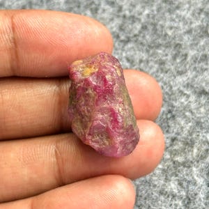 Könnte beinhalten: Ein roher, ungeschliffener Rubin-Edelstein mit tiefroter Farbe, akzentuiert durch hellere Rosa- und Gelbtöne. Der Naturstein hat eine strukturierte Oberfläche und eine unregelmäßige Form, die seinen rohen Zustand zeigt.