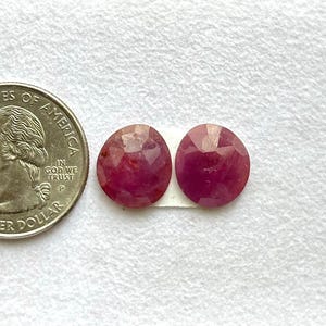 Puede incluir: Dos gemas facetadas, de forma ovalada y de color rojo intenso, se muestran junto a una moneda de 25 centavos estadounidense. Las gemas tienen una superficie pulida y reflectante.