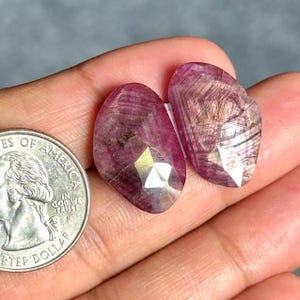 Könnte beinhalten: Zwei facettierte, oval geformte, tief magentafarbene Edelsteine mit sichtbaren inneren Einschlüssen und geometrischen Mustern. Eine silberfarbene US-Vierteldollarmünze liegt daneben, um die relative Größe der Steine zu verdeutlichen.
